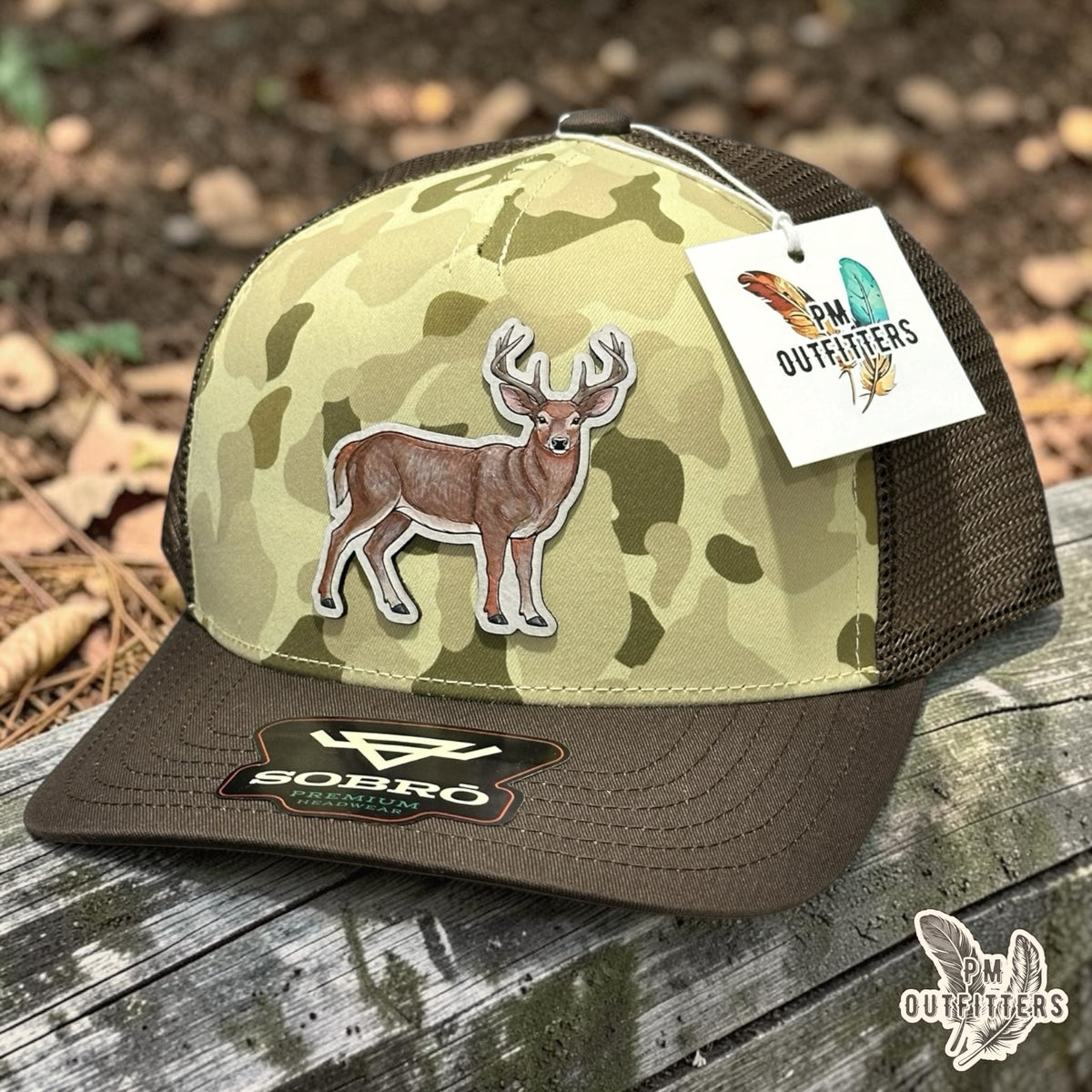 Hand-painted whitetail buck patch on a Sobro Waterwood 5-panel trucker hat in Ranchwood Camo with brown mesh back and bill, PM Outfitters True Originals