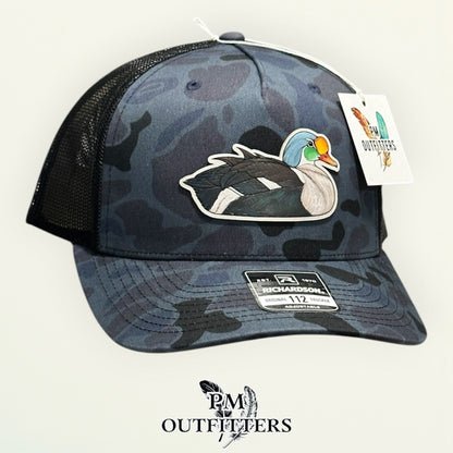Admiral duck camo Richardson 112PFP trucker hat with hand-painted King Eider patch, angled view, PM Outfitters True Originals