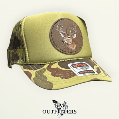 Hand-painted whitetail buck patch on a round brown vegan leather patch, olive green and camo Otto trucker hat, PM Outfitters True Originals